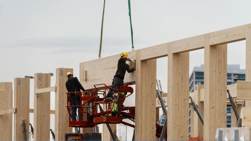 Zwei Arbeiter auf einer Hebebühne montieren sehr große Holzbau-Träger für ein großes Gebäude.