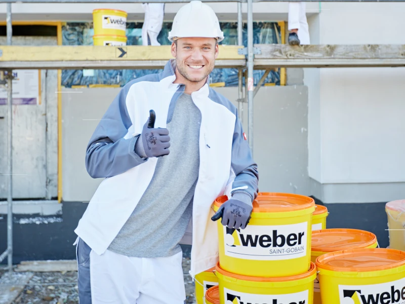 Ein Mann in weißer Arbeitskleidung lächelt mit einem erhobenen Daumen in das Bild. Sein Arm ist abgestützt auf den Eimern der Firma Saint-Gobain Weber.