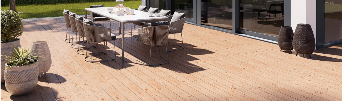 Eine große, sonnenbeschiene Holz-Terrasse vor einem modernen Holzbau mit einer großen Fensterfront. Auf der Terrasse steht ein großer Gartentisch und zehn Stühle.