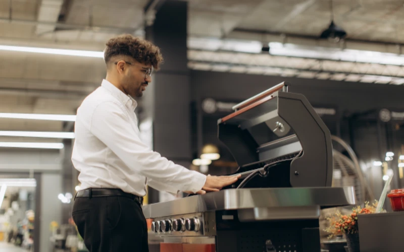 Das Bild zeigt eine Person in einem Verkaufsraum, die einen Gasgrill begutachtet. Der geöffnete Deckel und die Hand am Grillrost vermitteln eine typische Kaufsituation, bei der Ausstattung, Verarbeitung und Grillfläche geprüft werden.