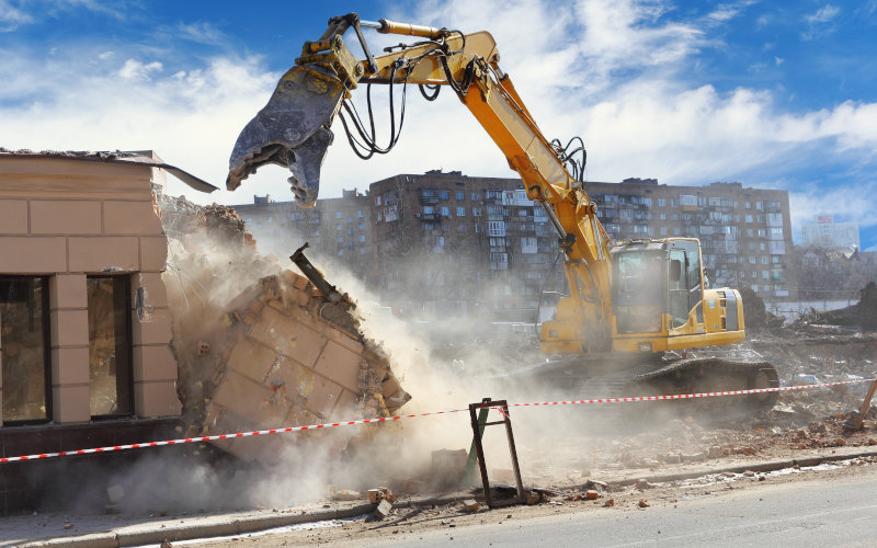 Ein Bagger reißt mit seiner Schaufel ein Gebäude ab, Staub und Trümmer fallen herunter.