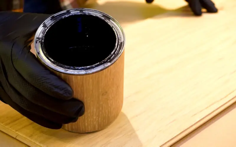 Hand mit Handschuh hält eine Dose Farbe über Holz.