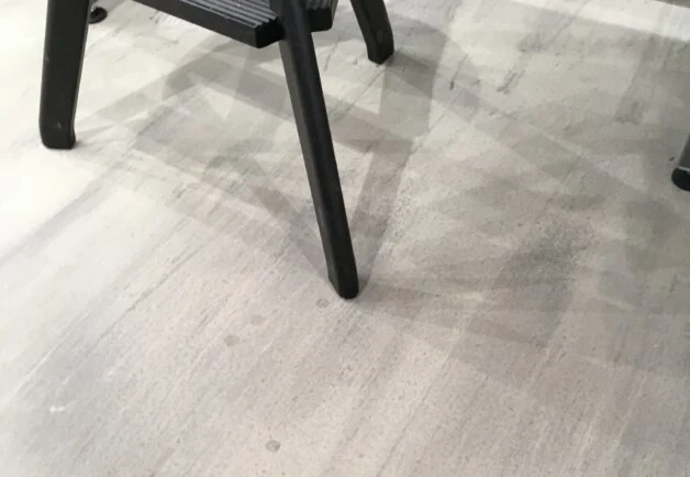 Ein Boden mit Flecken und Gebrauchsspuren. Auf dem Boden steht ein schwarzer Stuhl.