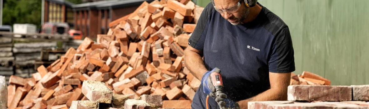 Handwerker sägt Holz zu. Im Hintergrund liegt ein Haufen mit roten Ziegelsteinen.