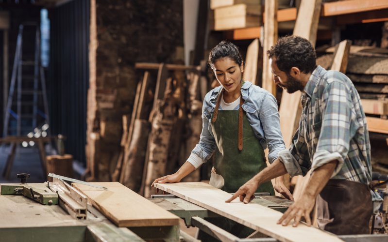 Tischler und Tischlerin unterhalten sich in einer Werkstatt über vorliegendes Holz