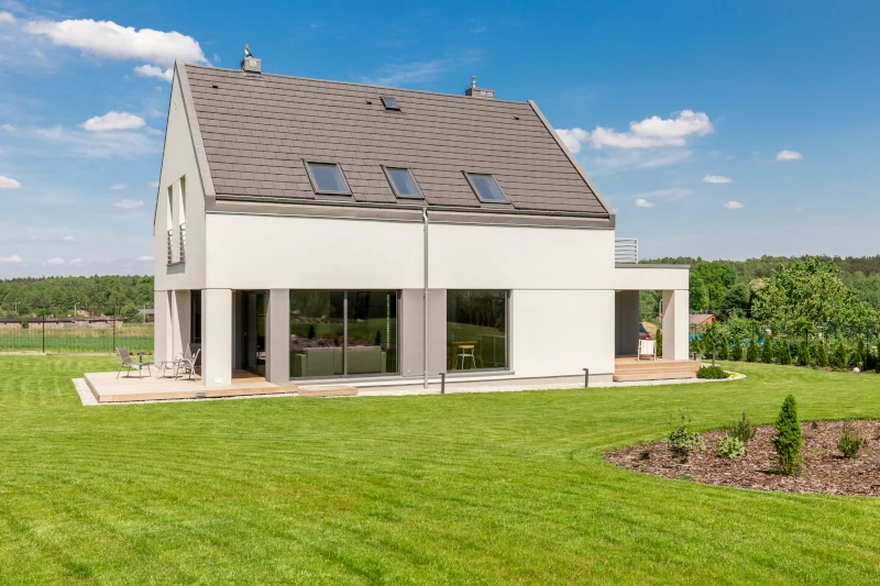 Zu sehen ist ein Haus mit einer großen Fensterfläche, stehend auf einem zwar grünen aber auch kargen Grundstück. Das Haus weißt außerdem eine kleine Holzterrasse auf.