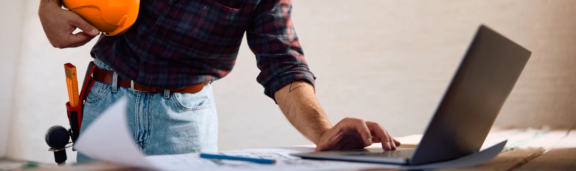 Ein Mann steht an einem Tisch, hält einen orangefarbenen Schutzhelm in der linken Hand und bedient mit der rechten Hand einen Laptop. Auf dem Tisch liegen Papiere und ein Stift. Er trägt ein kariertes Hemd und Jeans.