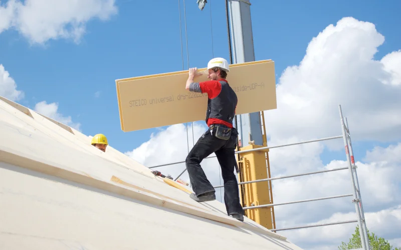 Dachdecker steigt auf das Dach mit Dämmplatte in der Hand, im Hintergrund ein Kran und Gerüst