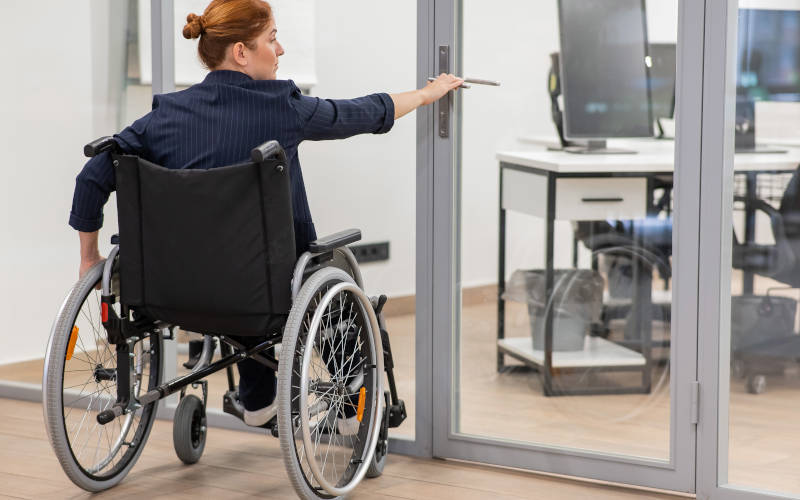Frau im Rollstuhl öffnet eine Glastür vor sich 