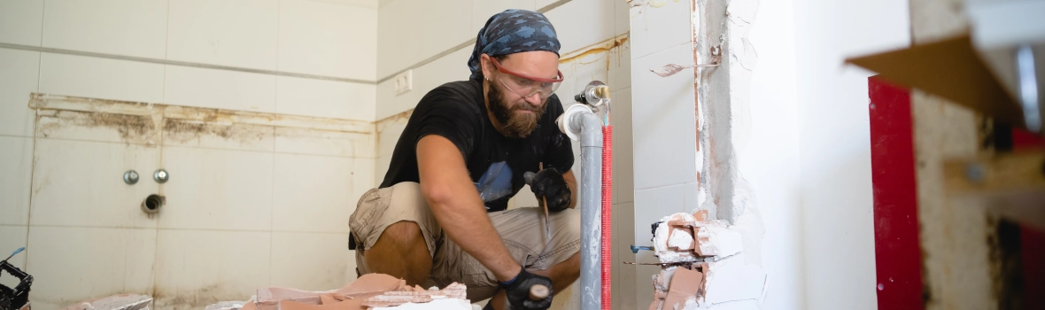 Ein Bauarbeiter, der ein Badezimmer renoviert, umgeben von Bauschutt und zerbrochenen Fliesen, zeigt Konzentration und Geschick beim Ausbau alter Armaturen mit Werkzeugen