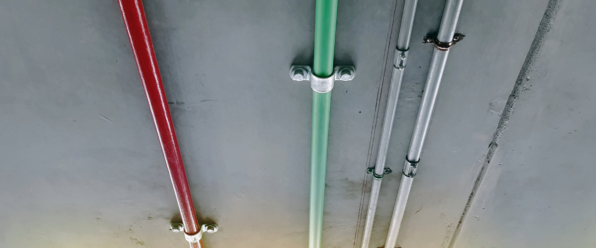 Mehrere Rohre in verschiedenen Farben (rot, grün, grau) verlaufen parallel zueinander an einer grauen Decke.