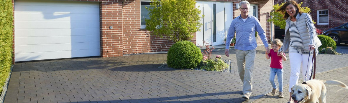 Mann und Frau laufen mit kleinem Kind an der Hand und Hund die Auffahrt vor ihrem Haus mit Garagentor und Haustür entlang