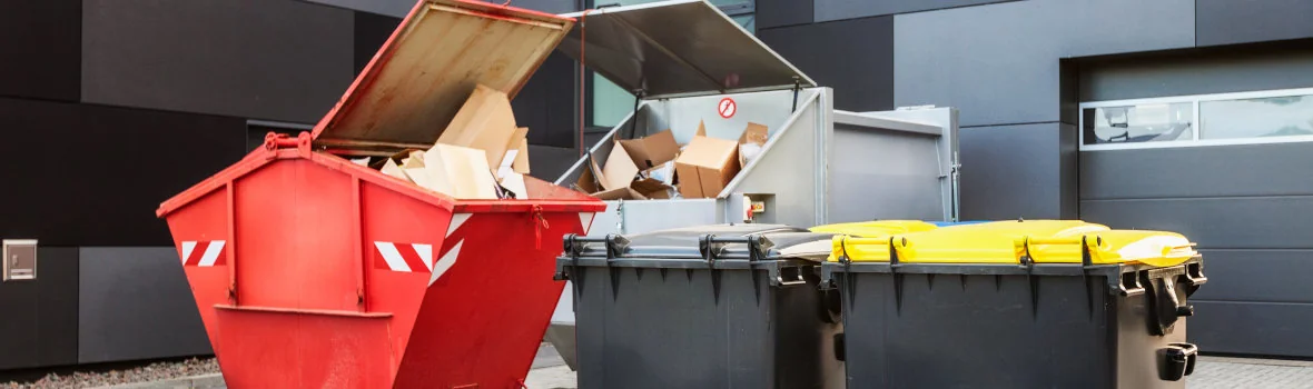 Mehrere Mülltonnen und gefüllte Container stehen vor einem modernen Gebäude