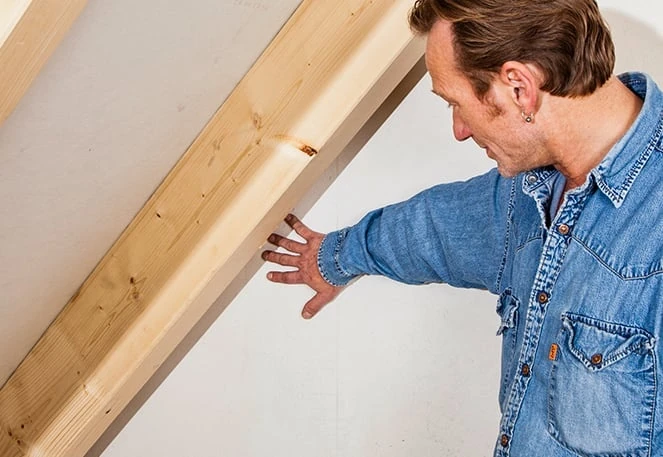 Das Bild zeigt eine Person, die eine Wand oder ein Bauteil in einem Dachgeschoss inspiziert. Die Person trägt ein Jeanshemd und hat eine Hand auf das Bauteil gelegt.