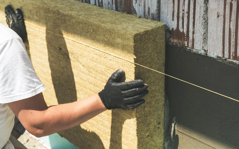 Person installiert Dämmplatte an Wand, trägt Handschuhe, verwendet Schnur für präzise Ausrichtung.