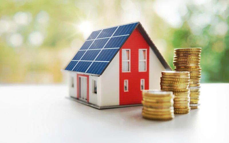 Miniaturhaus mit Solarpaneelen neben Stapeln von Münzen.