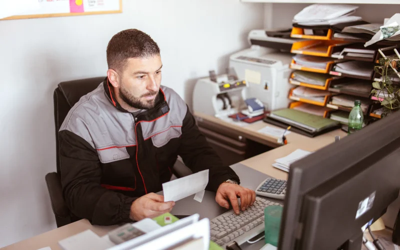 Ein Mann der in einem Büro sitzt und auf einer Tastatur tippt. In der rechten Hand befindet sich ein Zettel. Im Hintergrund befinden sich übliche Bürointensilien wie ein Locher und Zettelablagen.