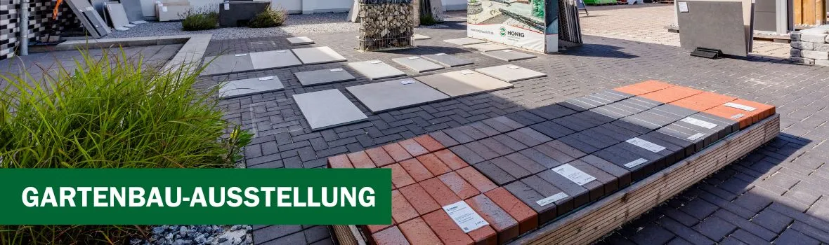 Titelbild der Gartenbau-Ausstellung der HONIG Baustoffe Website. Es ist ein Teil der Außenausstellung zu sehen mit verschiedenen Kopfsteinpflastern, die mit Beschreibungen versehen sind. Links ist ein Gebüsch, das Wetter ist sonnig.