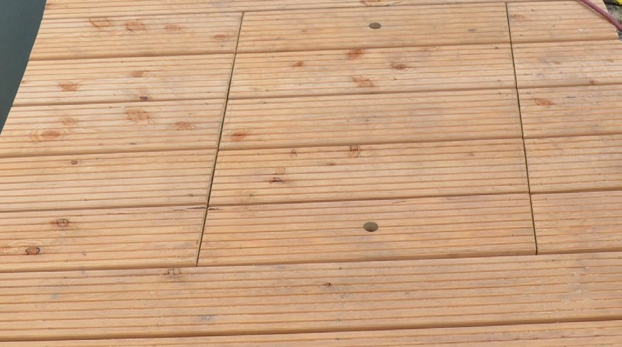 Nahaufnahme von hellen Holzplanken mit parallelen Rillen. In den Planken ist ein quadratischer Zugangspunkt mit zwei runden Grifföffnungen zu sehen.
