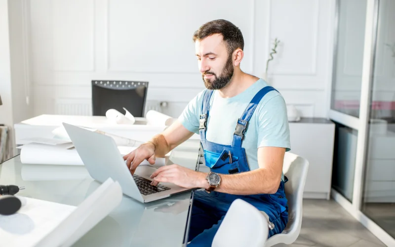 Handwerker sitzt mit Laptop an einem Tisch umgeben von Dokumenten