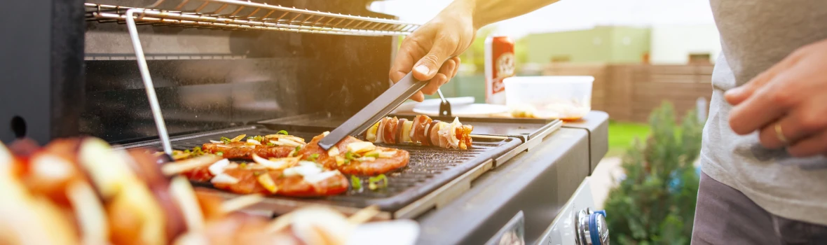 Das Bild zeigt eine Person beim Grillen an einem Gasgrill im Garten oder auf der Terrasse. Auf der Grillplatte liegen verschiedene Speisen, während die warme Lichtstimmung eine sommerliche, entspannte Grillatmosphäre vermittelt.