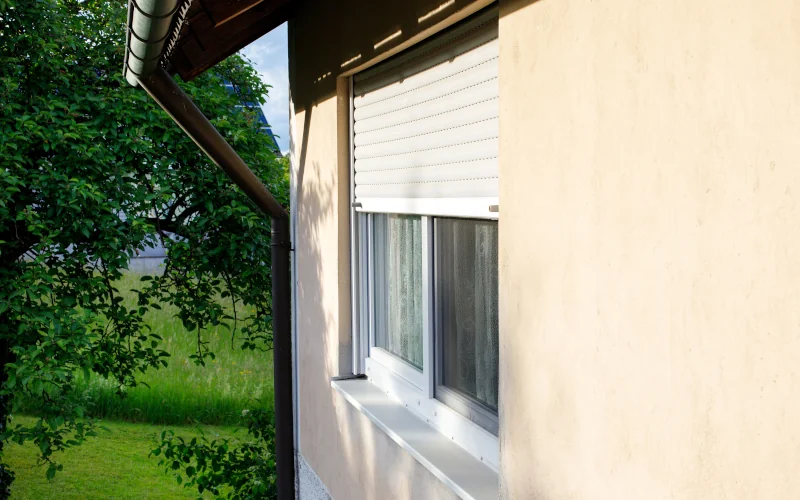 Haus mit beigefarbener Fassade mit halb heruntergelassenem Rollladen angestrahlt von der Sonne