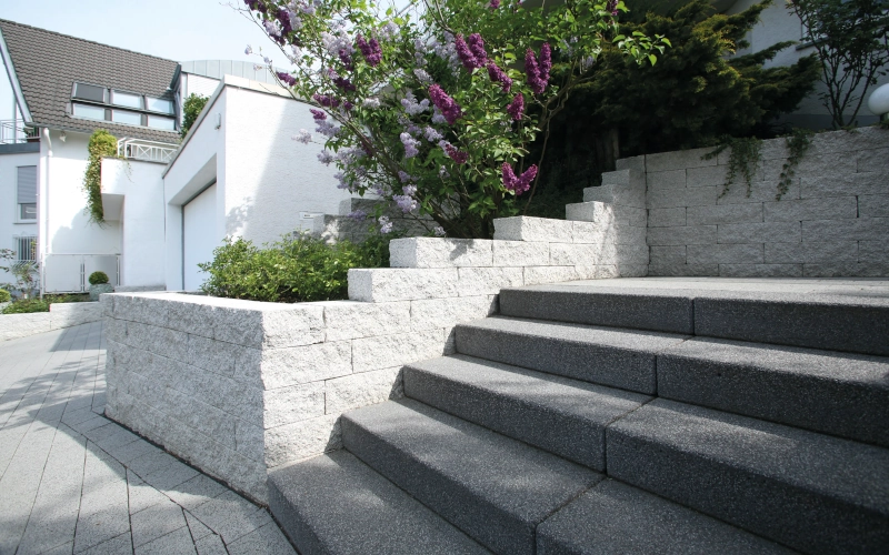 Weiße mauer mit grauer Treppe, mit einem Einfamilienhaus im Hintergrund.