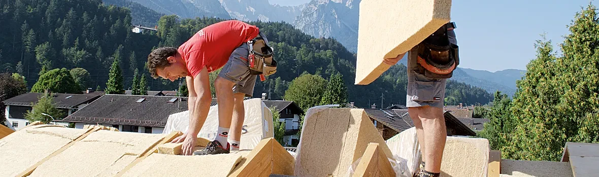 Zwei Männer stehen auf einem ungedecktem Hausdach. Einer von ihnen trägt ein Stück Dämmstoff. Der andere Mann verlegt den Dämmstoff auf dem Dach. 