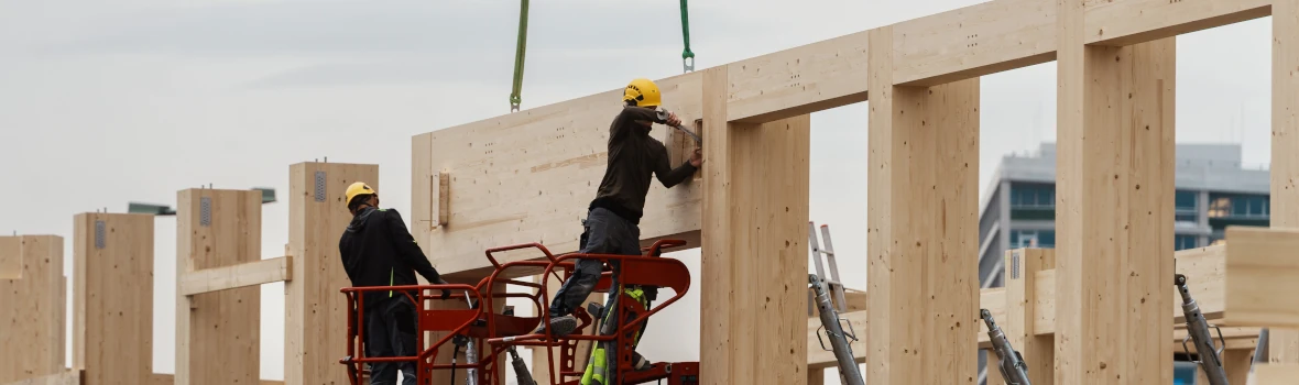 Zwei Arbeiter auf einem Hubgerüst bauen ein großes Holzständerwerk zusammen.