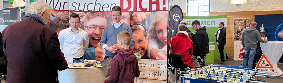 Eine Jobmesse zeigt verschiedene Stände, darunter ein Tischfußballspiel und Werbeplakate für Berufsausbildung.