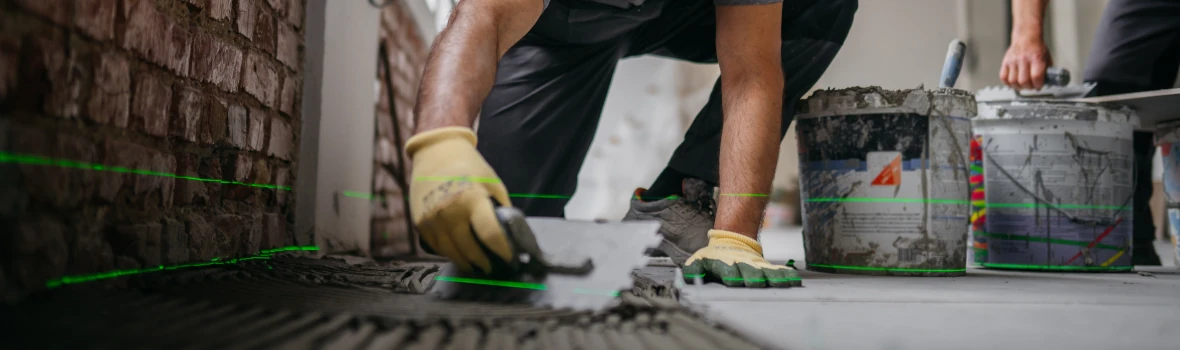 Das Bild zeigt eine Person, die mit Handschuhen auf dem Boden arbeitet. Sie trägt Mörtel mit einer Kelle auf. Im Hintergrund sind ein Eimer und Werkzeuge zu sehen, außerdem verlaufen grüne Laserlinien über Boden und Wand.