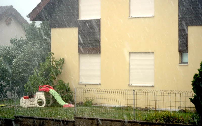 gelbes Haus mit geschlossenen Außenrollläden bei Starkregen und Hagel