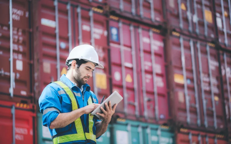 Arbeiter steht mit Tablet vor einer Wand aus Containern 