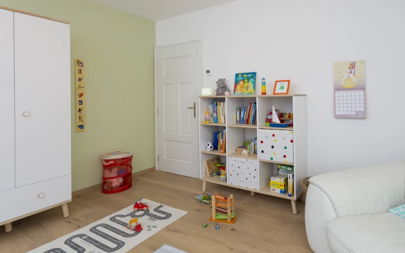 Das Bild zeigt ein helles Kinderzimmer mit weißem Kleiderschrank, Spielteppich, Bücherregal, Spielzeug und einer gemütlichen Sitzecke.