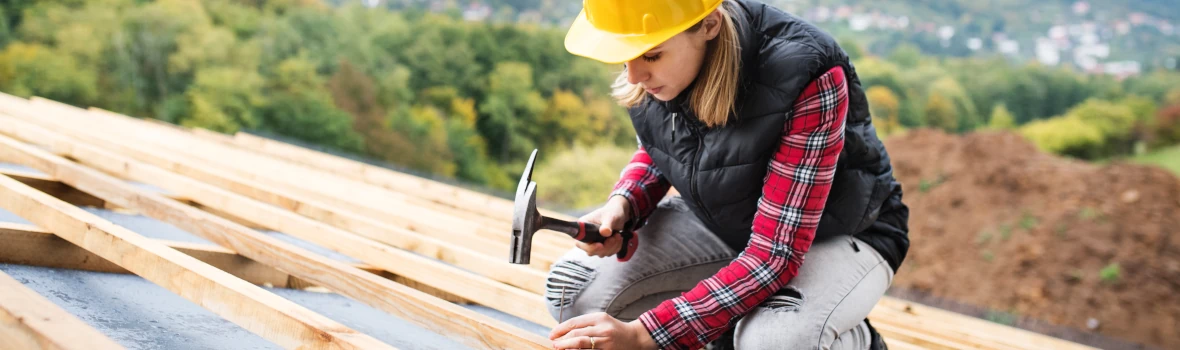 Eine Dachdeckerin mit gelbem Helm und Hammer kniet arbeitend auf der Lattung eines neuen Daches.