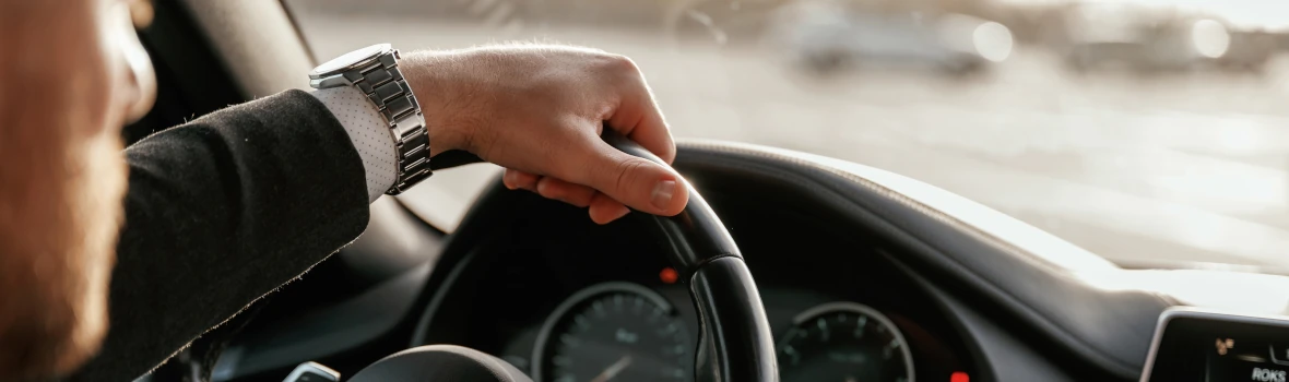 Das Bild zeigt eine Person, die am Steuer eines Autos sitzt und das Lenkrad mit beiden Händen hält. Sichtbar sind das Armaturenbrett, die Instrumente und ein Teil des Innenraums.