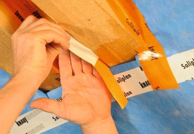 Eine Hand, die ein Stück Klebeband abzieht und es auf eine blaue und orangefarbene Oberfläche aufbringt. Das Klebeband trägt die Aufschrift "Knauf Soliplan".