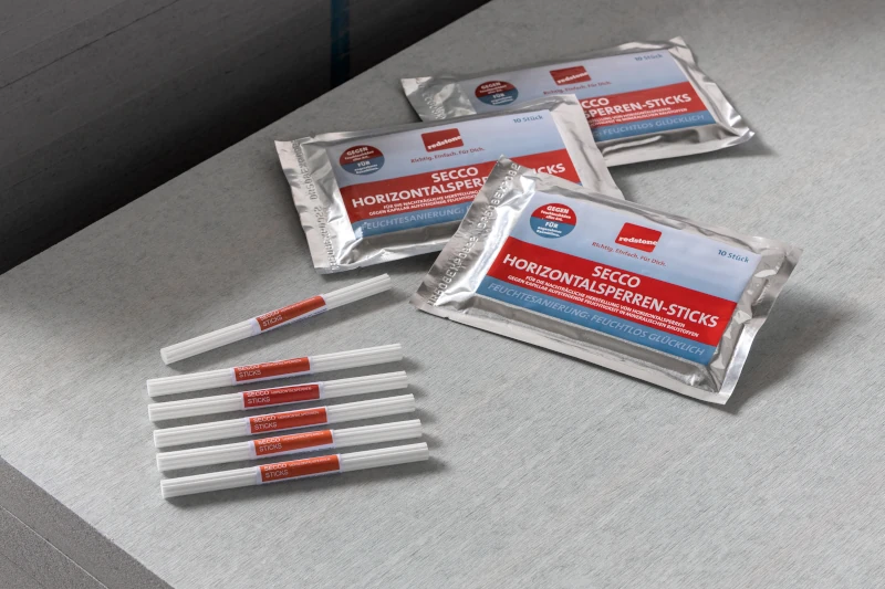 Drei geschlossene Packungen Secco Horizontalsperren-Sticks liegen auf dem Tisch. Sechs Sticks liegen ebenfalls zur Veranschaulichung auf dem Tisch.