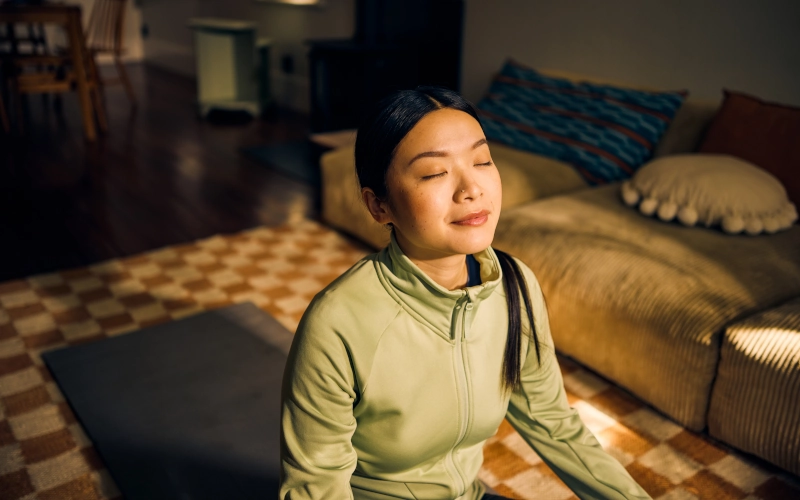 Junge asiatische Frau meditiert im Wohnzimmer. Indoor-Lifestyle-Porträt. Selbstfürsorge- und Achtsamkeitskonzept. 