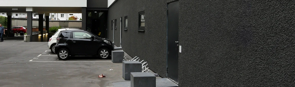 Eine schwarze, gedämmte Wand mit mehreren Fenstern und einer schwarzen Tür ist abgebildet. Vor dem Gebäude befindet sich eine Parkfläche und Fahrradständer. 