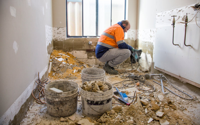 Ein Bauarbeiter renoviert mit Werkzeugen ein Zimmer, das voller Schutt und Baumaterialien ist.