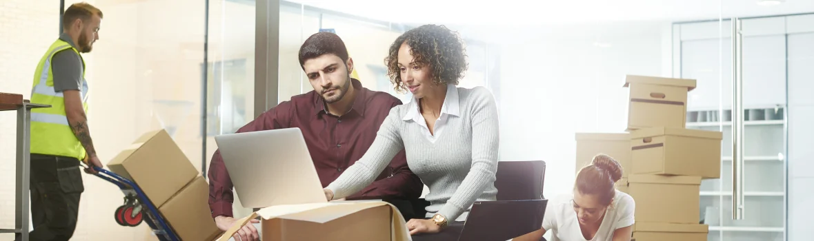 Ein Mitarbeiter und eine Mitarbeiterin packen Umzugskartons im Büro ein. Im Hintergrund läuft ein Mann mit Warnweste und schiebt Kartons auf einer Sackkarre.
