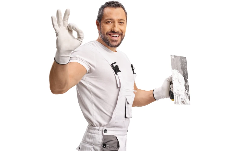 Das Bild zeigt einen lächelnden Handwerker in heller Arbeitskleidung, der eine Kelle mit Putz in der Hand hält und mit der anderen Hand ein Okay-Zeichen macht.