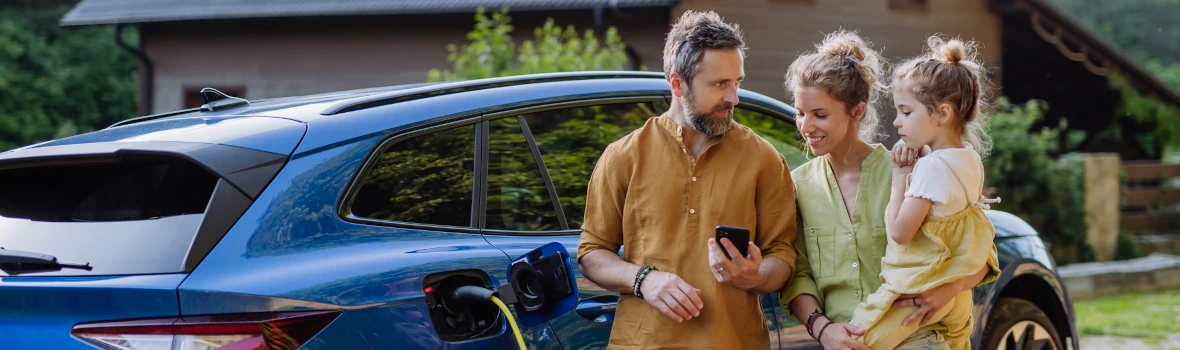 eine dreiköpfige Familie steht vor einem ladenden blauen E-Auto vor einem Haus 