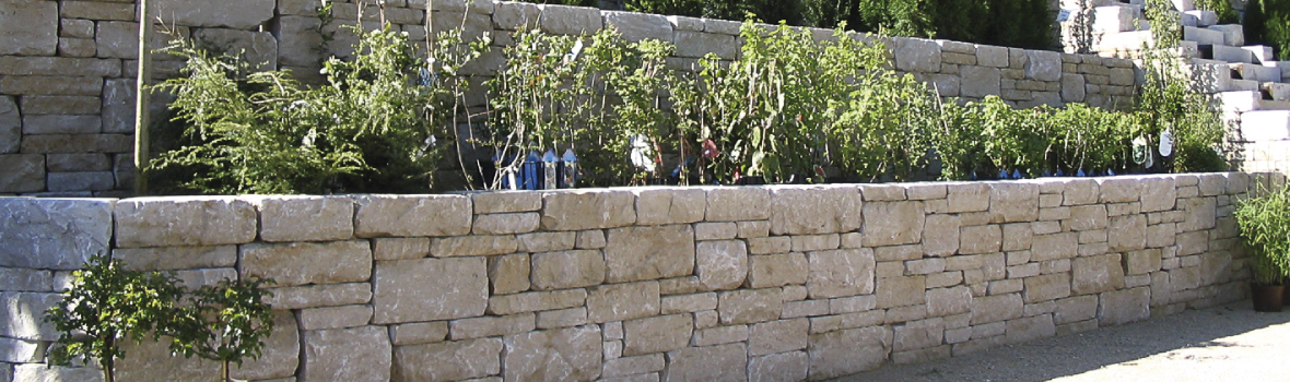 Mehrstufige Trockenmauern aus hellen, unregelmäßig geformten Natursteinen stützen bepflanzte Terrassenbeete. In den Beeten wachsen dichte grüne Pflanzen und einige Tomatenstauden. Rechts führen Natursteinstufen nach oben. Der Bereich ist sonnig und wirkt gepflegt.
