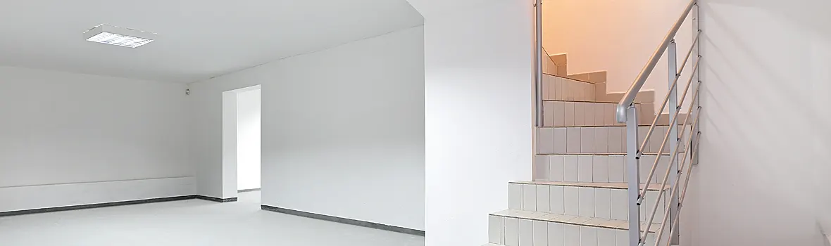 Leerer, weißer Kellerraum mit Türöffnung und angrenzender Treppe mit Geländer.