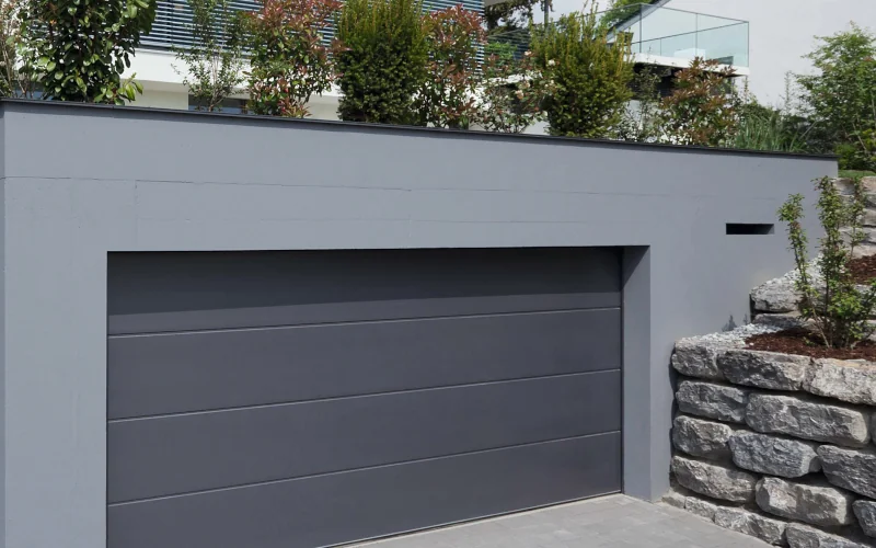 graue Garage mit grauem Tor on vielen Pflanzen umgeben 