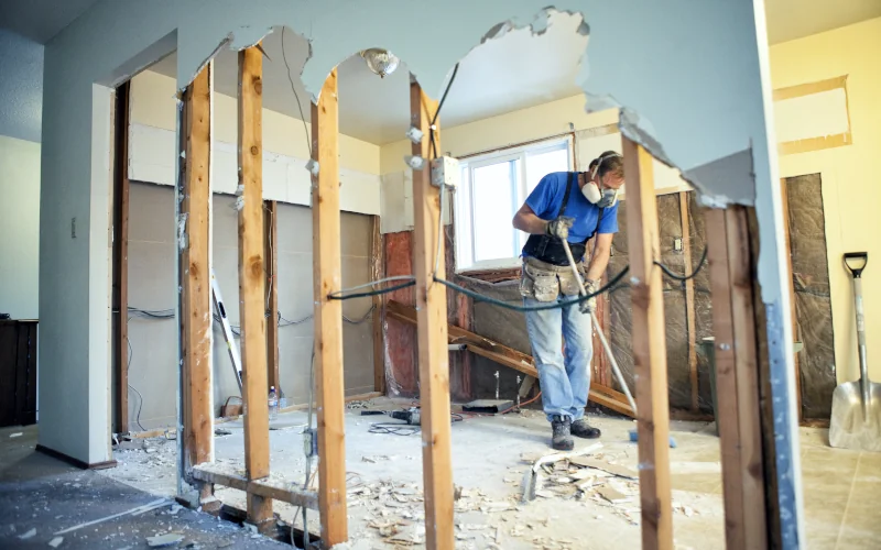 Hausrenovierung, Arbeiter entfernt Wand, trägt Maske und blaue Kleidung.