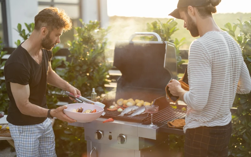 Das Bild zeigt zwei Personen beim Grillen auf einer Terrasse oder im Garten. Auf dem Gasgrill liegen verschiedene Speisen, während die warme Abendstimmung eine entspannte, gesellige Grillsituation vermittelt.