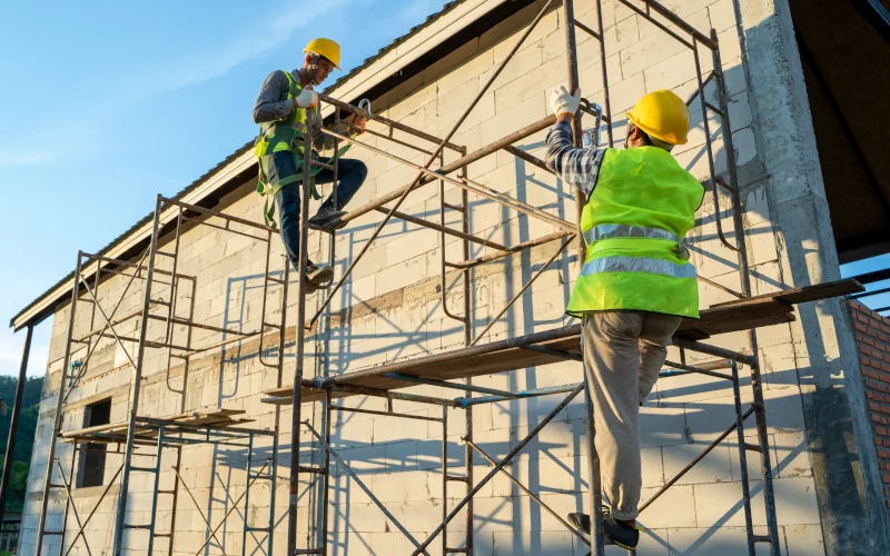 Bauarbeiter auf Gerüst, tragen Schutzhelme und Westen, arbeiten an Gebäude, sonniger Tag.